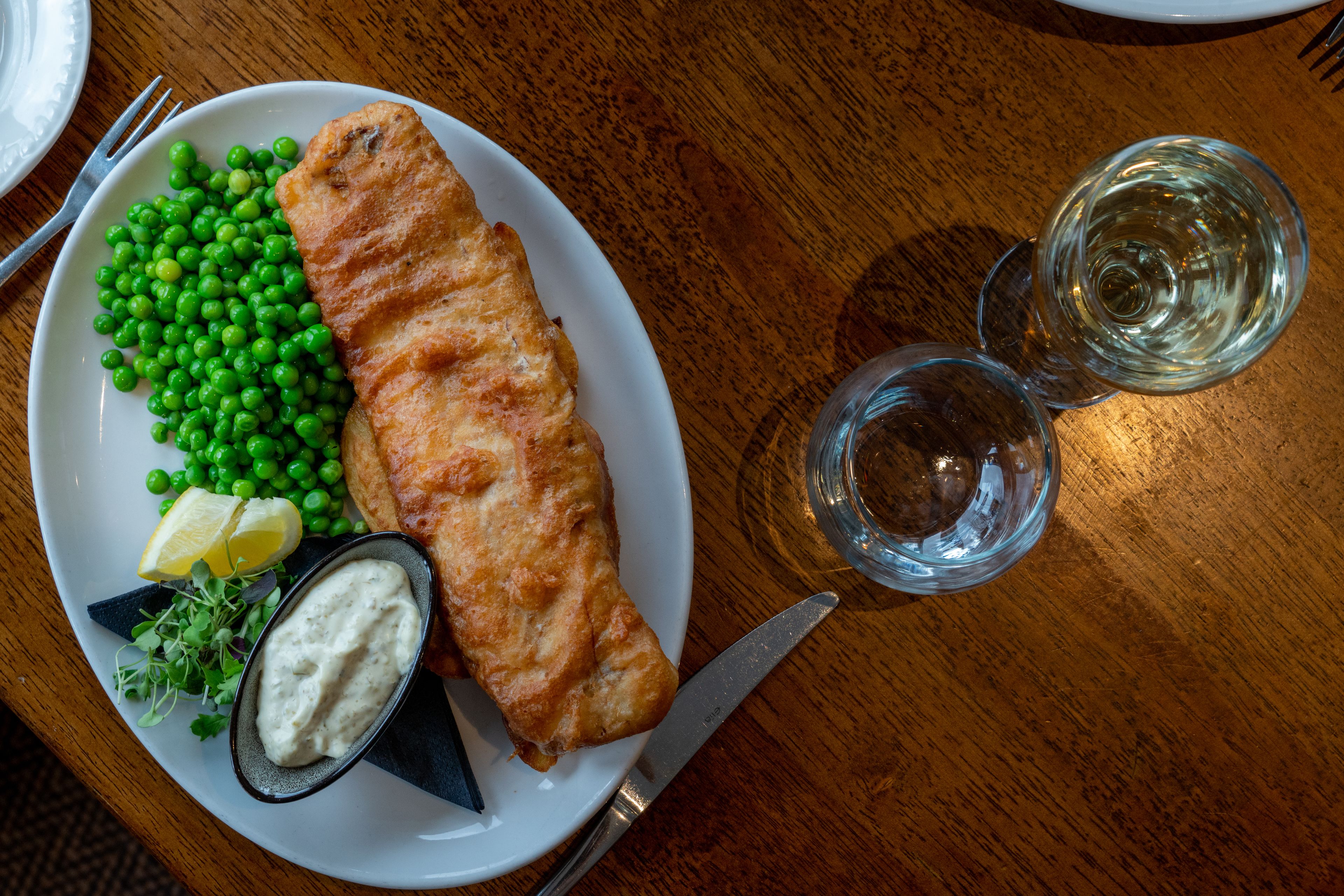 A plate with a fish fillet, peas, a lemon wedge, and a small bowl of sauce on a wooden table.
