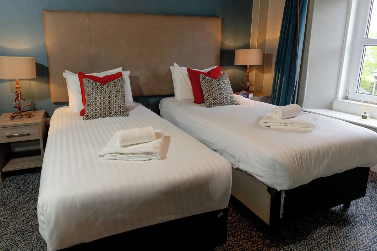 A hotel room with two beds, a nightstand with a lamp, and a window with curtains.