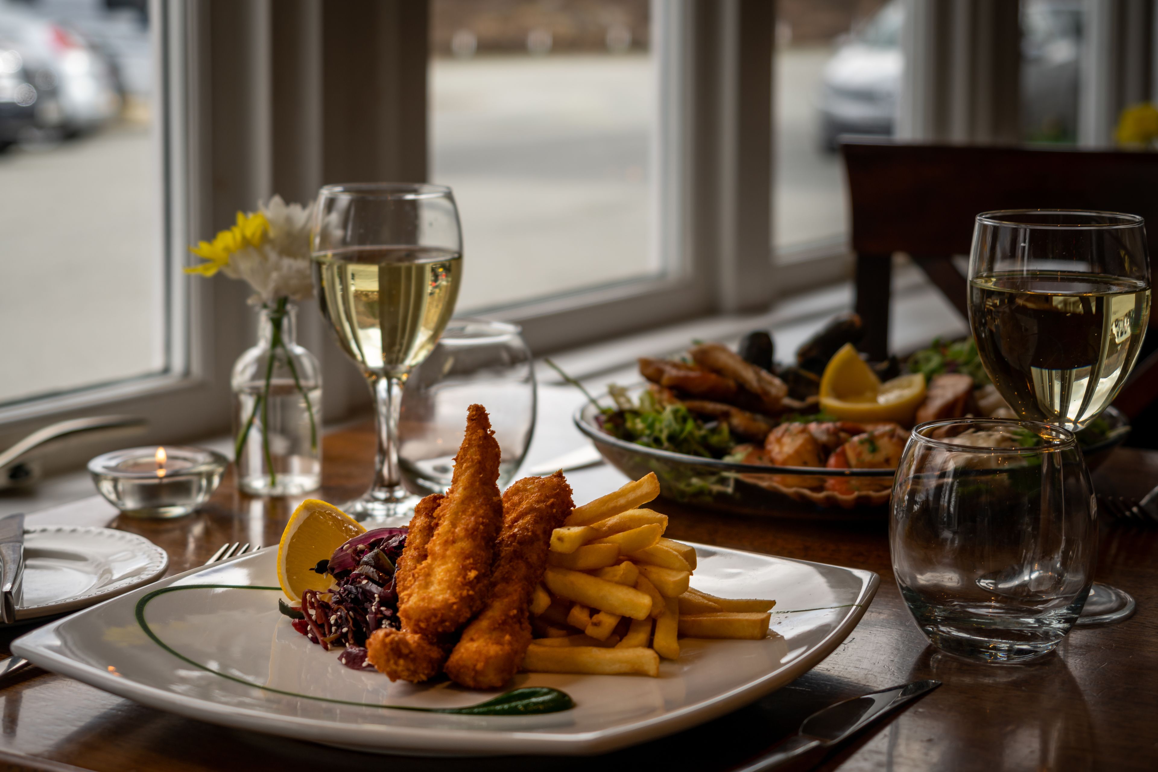 A dining table with a plate of fish and chips, a glass of white wine, and a vase with flowers.