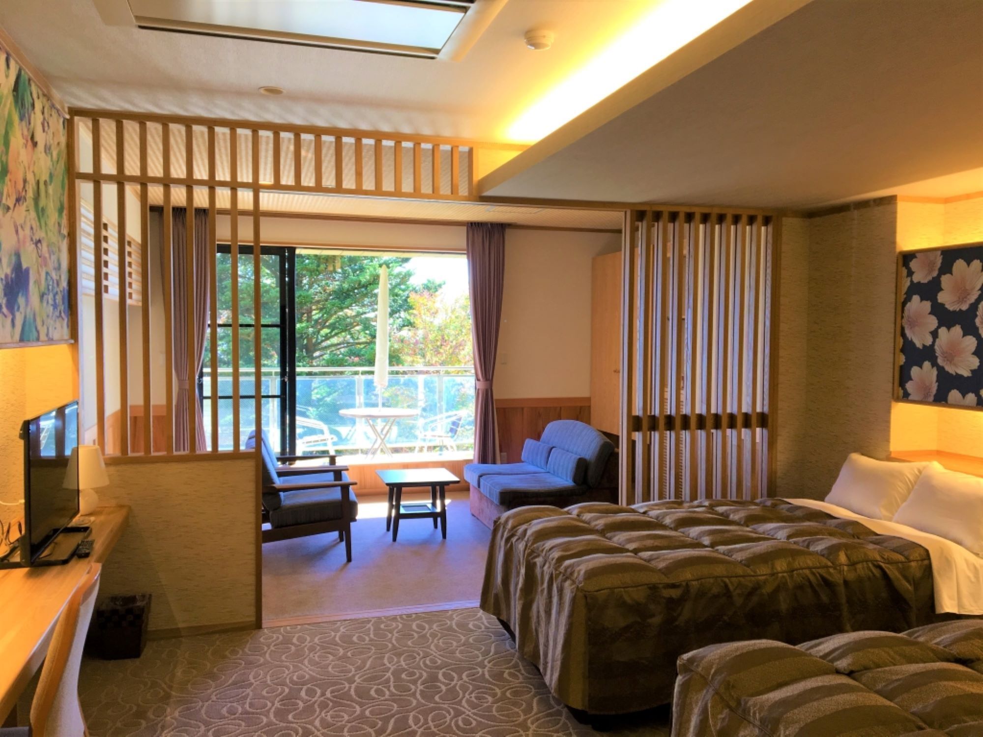 Quarto de hotel com cama, escrivaninha, cadeira, mesa pequena e varanda com vista para árvores.
