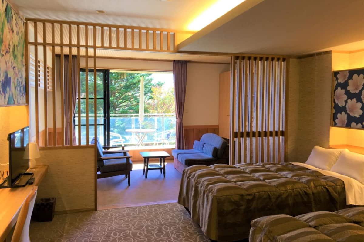 A hotel room with a bed, a desk, a chair, a small table, and a balcony with a view of trees.
