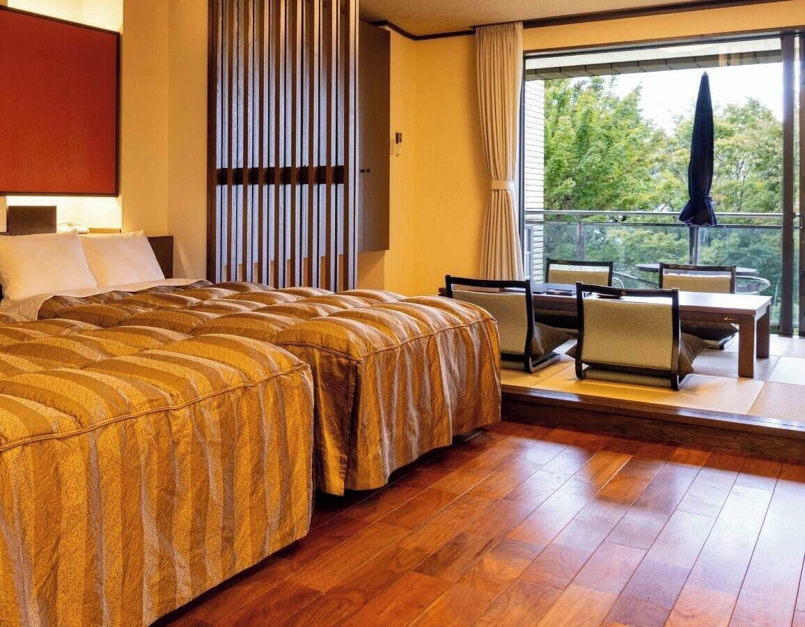 Quarto de hotel com uma cama grande, área de estar com mesa e cadeiras, janela com vista para árvores e cortina.