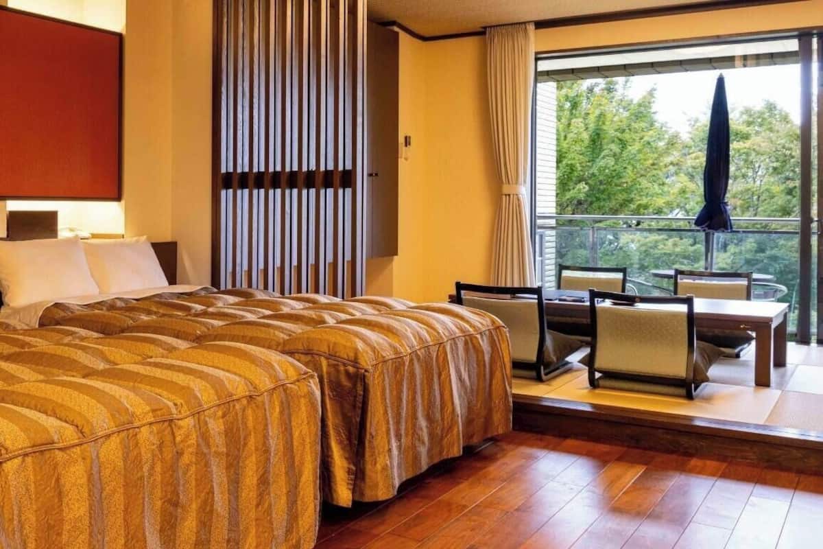A hotel room with a large bed, a seating area with a table and chairs, a window with a view of trees, and a curtain.