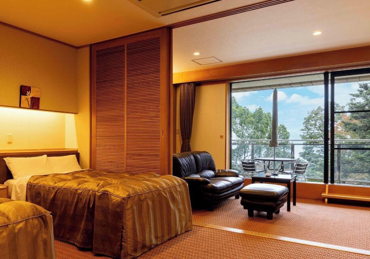 A hotel room with a bed, a sofa, an ottoman, a balcony with a table and chairs, and a large window.