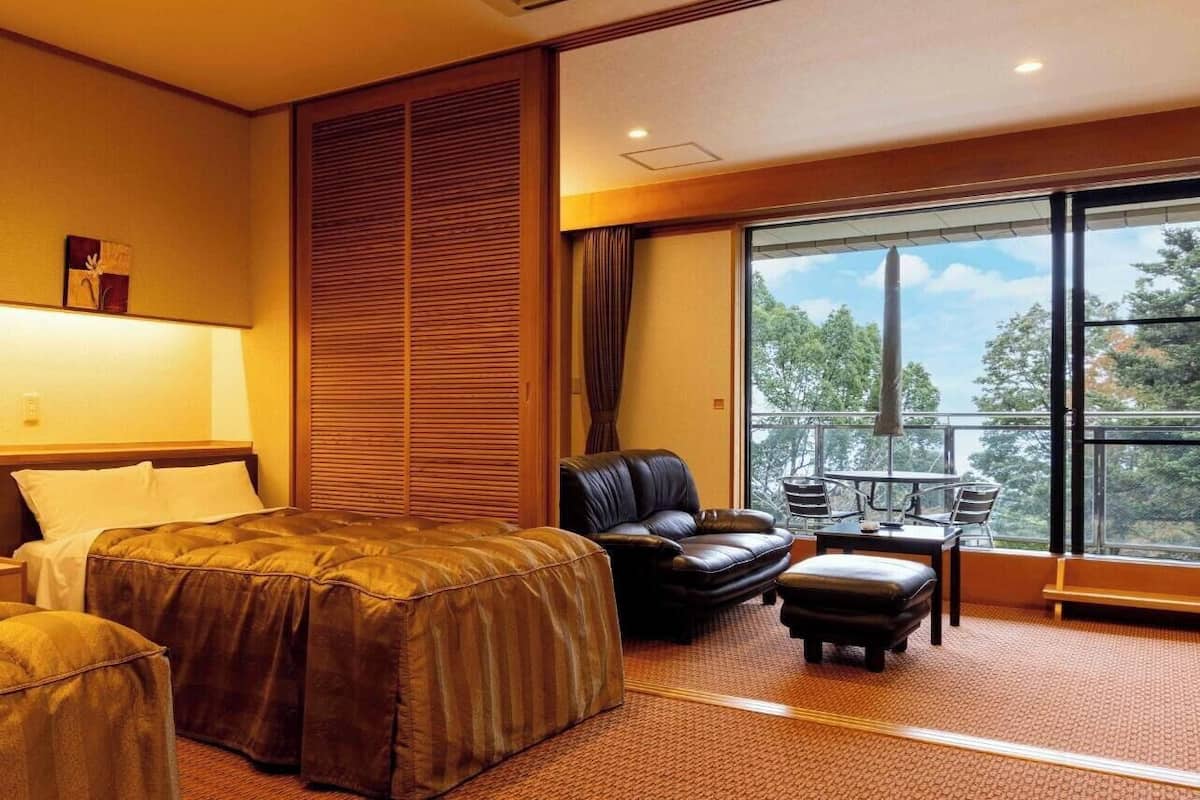 A hotel room with a bed, a sofa, an ottoman, a balcony with a table and chairs, and a large window.