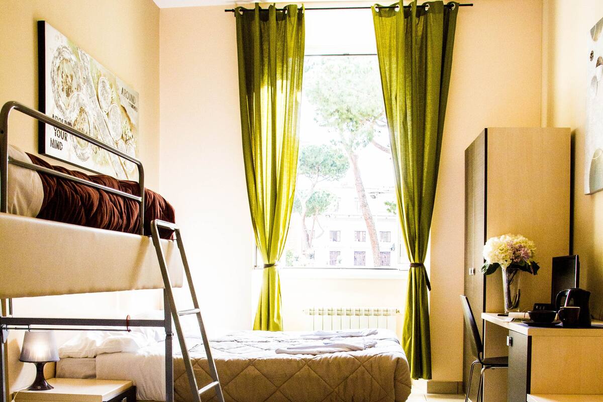 A dorm-style room with bunk beds, a desk, a chair, and a window with curtains.