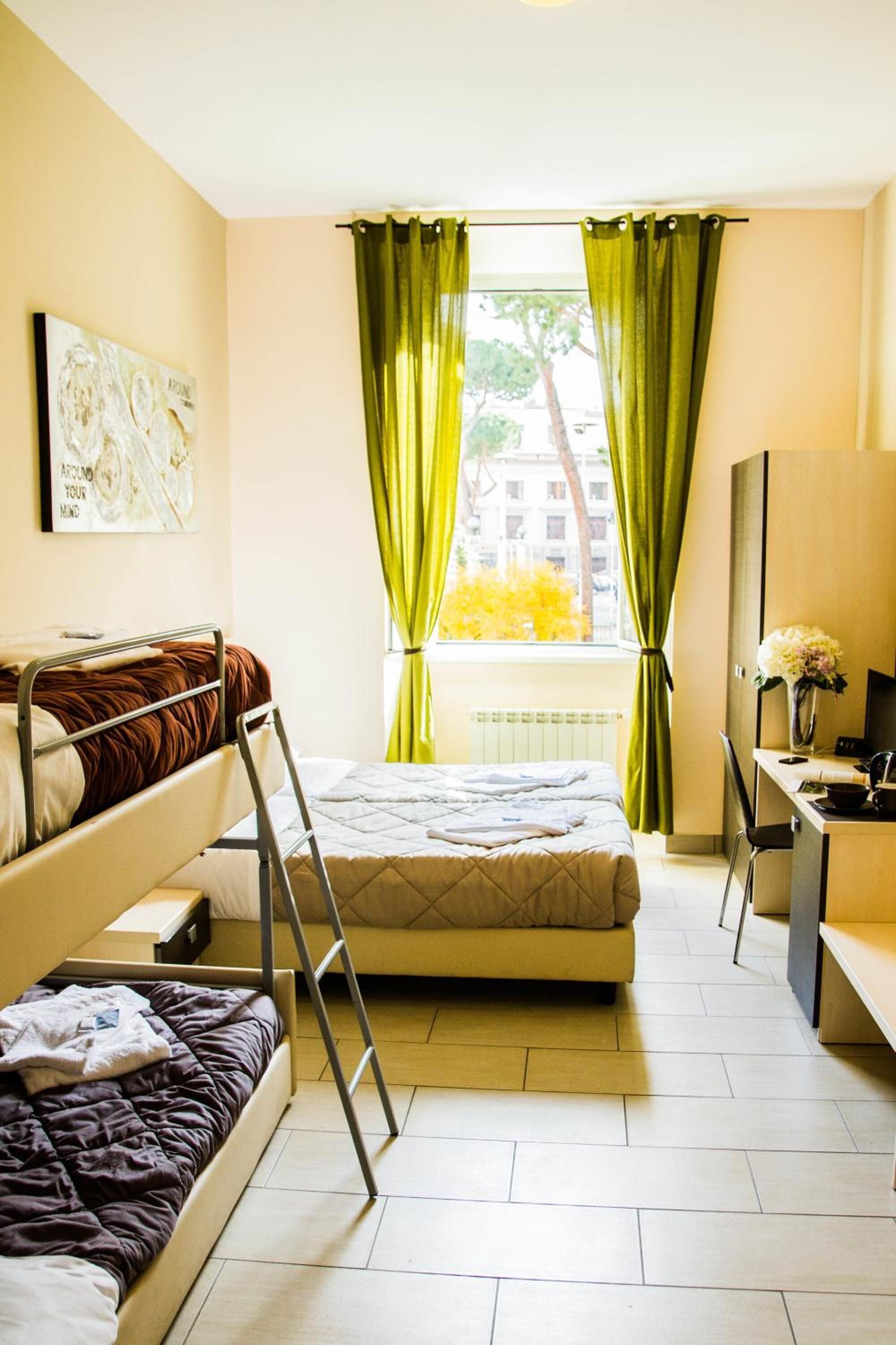 A compact hotel room with a bunk bed, a desk with a chair, a mirror, and a window with curtains.