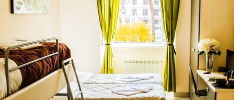 A compact hotel room with a bunk bed, a desk with a chair, a mirror, and a window with curtains.