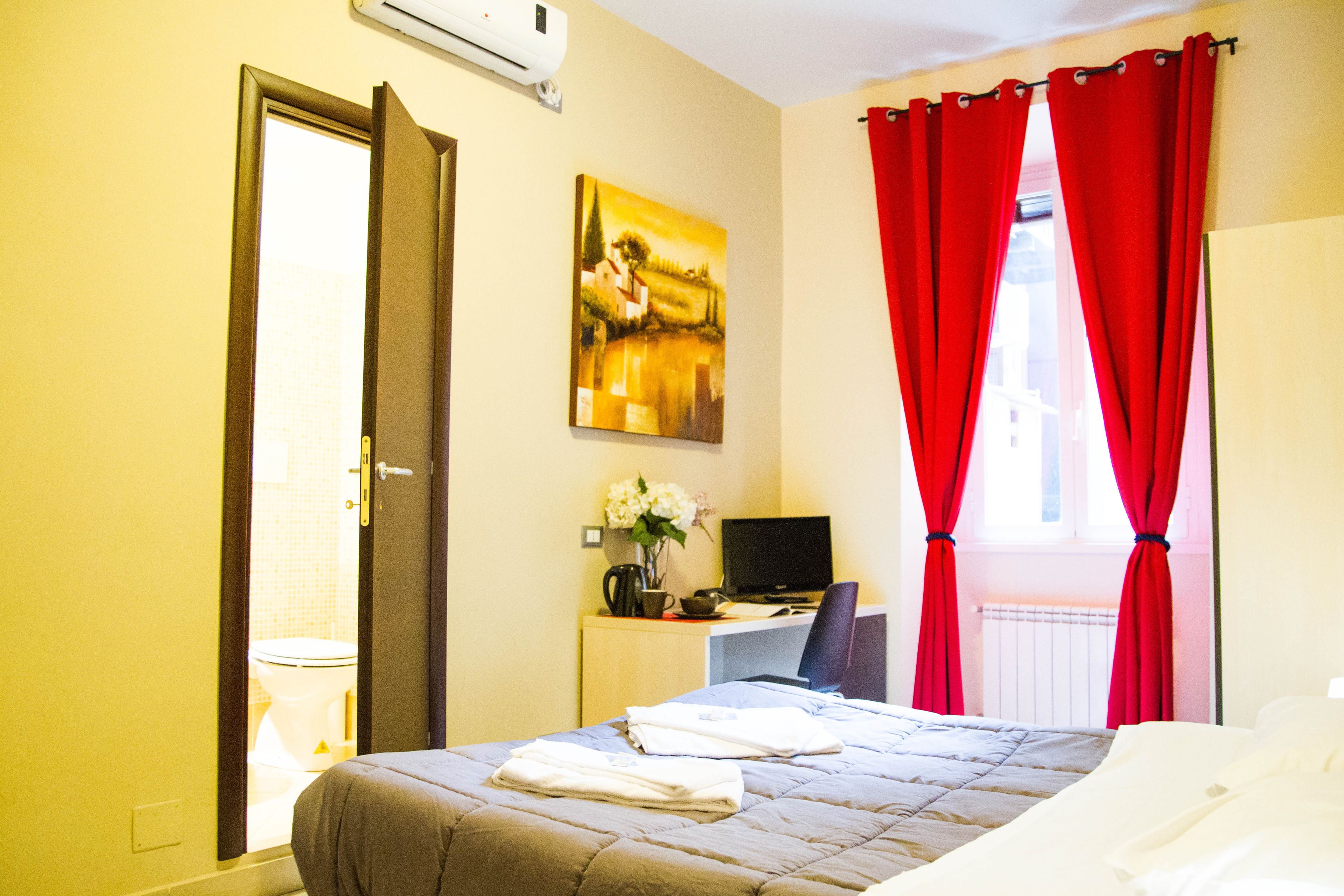 A hotel room with a bed, a desk with a computer, a chair, and a window with red curtains.