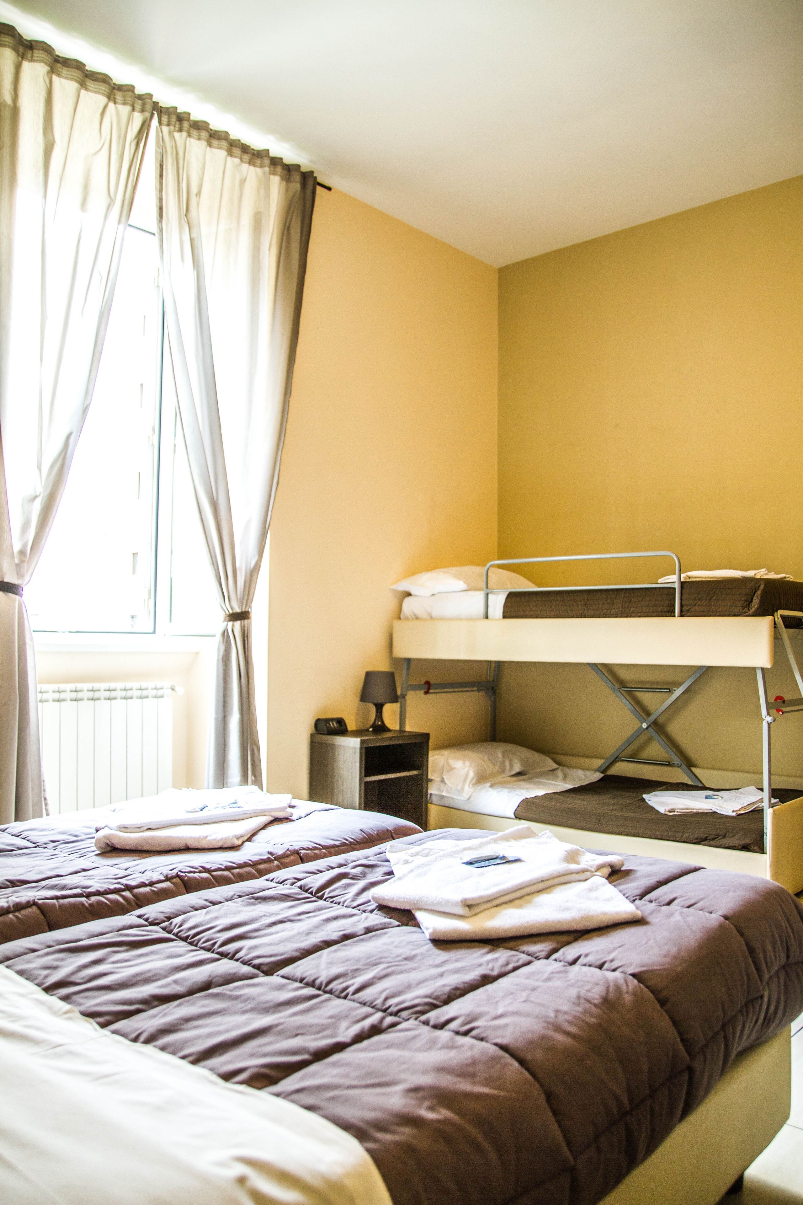 A room with a bunk bed, a single bed, a radiator, a nightstand with a lamp, and a window with curtains.