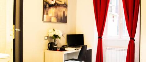 A hotel room with a bed, a desk with a computer, a chair, and a window with red curtains.