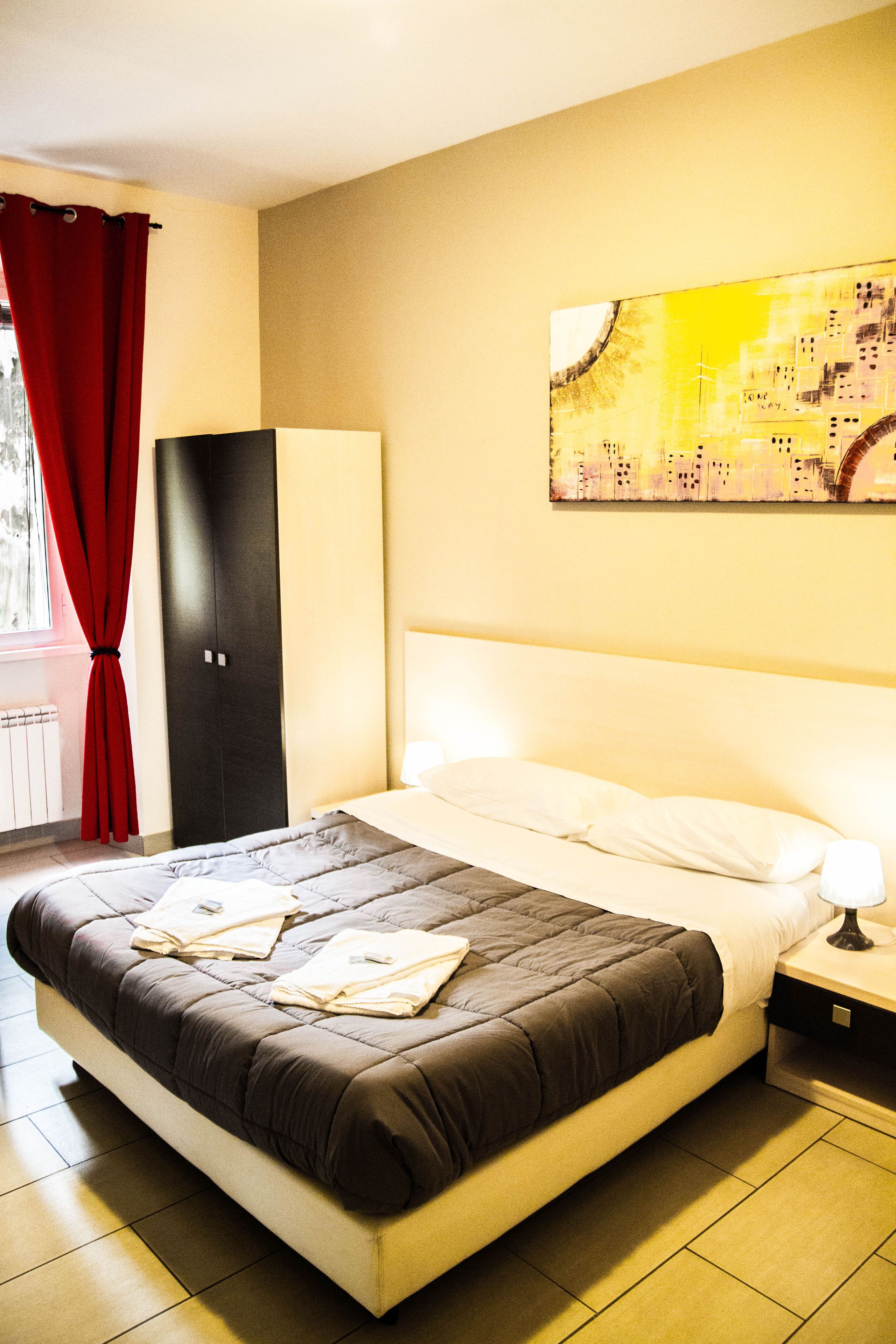 A hotel room with a bed, a nightstand, a red curtain, a black wardrobe, and a cityscape painting on the wall.