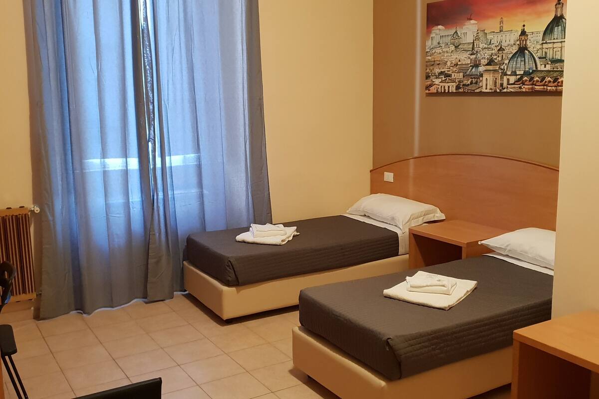 A hotel room with two beds, a radiator, a window with curtains, and a cityscape painting on the wall.