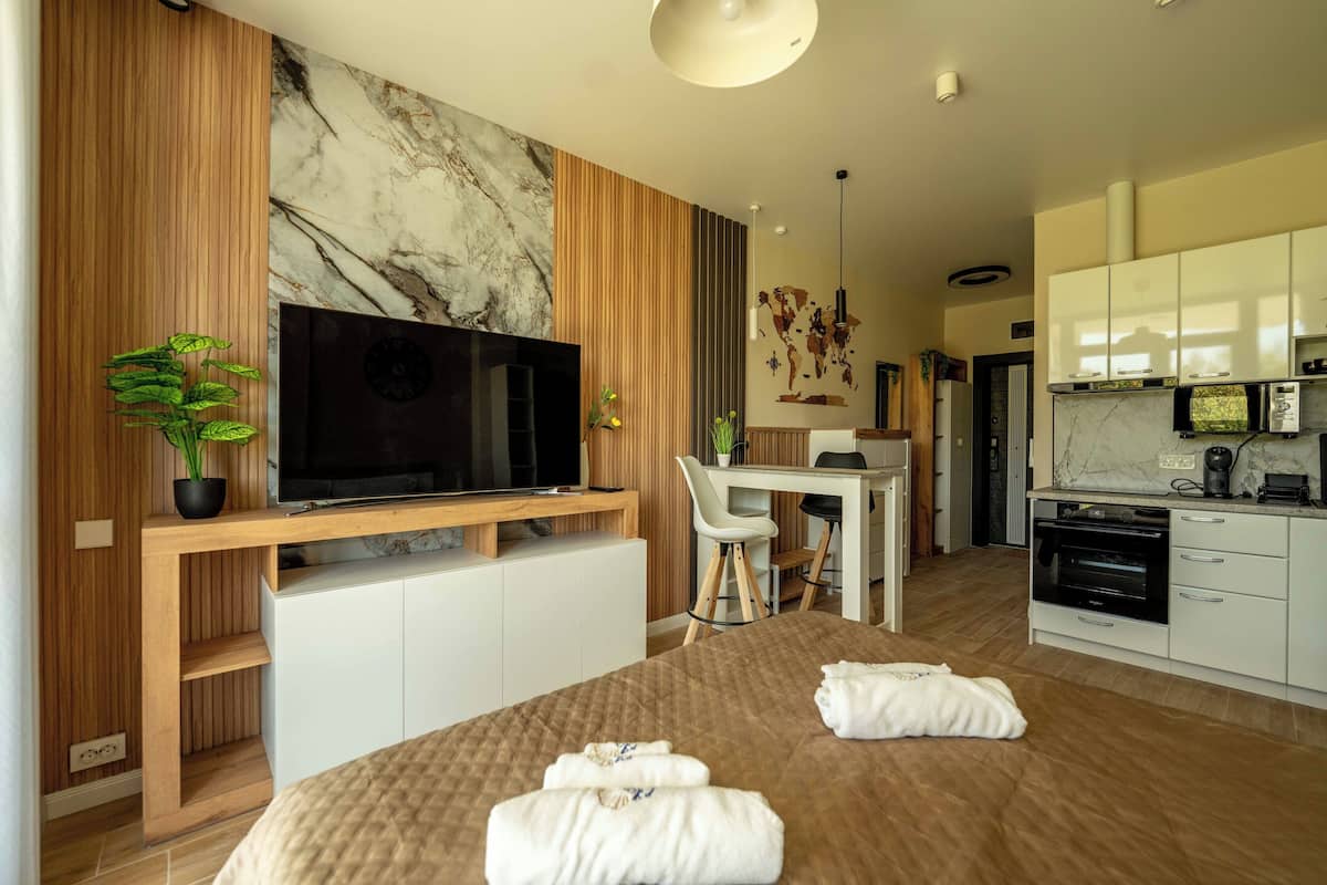 A modern hotel room with a large bed, a TV, a desk, and a kitchen area.
