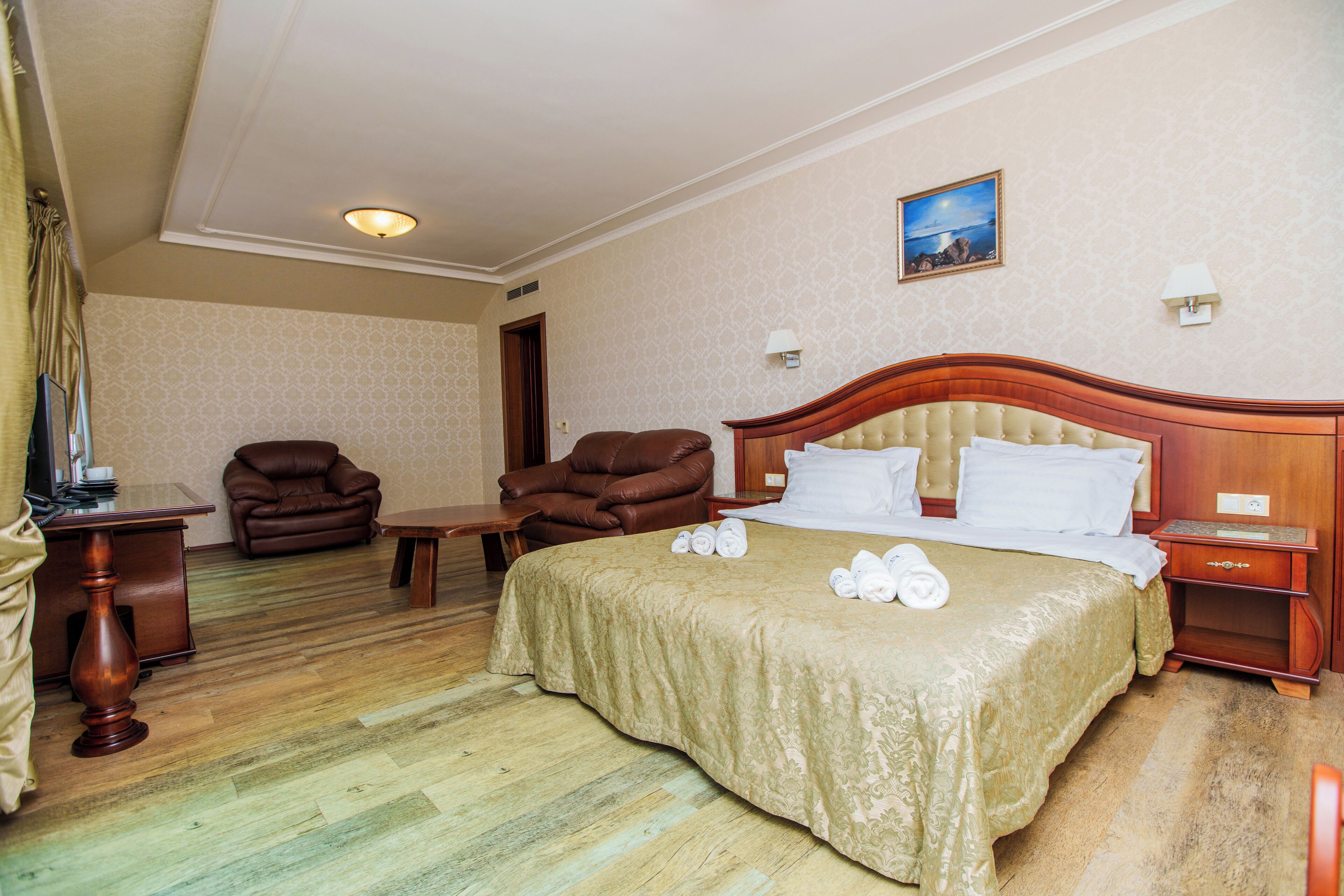 Habitación de hotel con una cama grande, cabecera de madera, un sillón de cuero marrón, una mesita y un escritorio con lámpara.