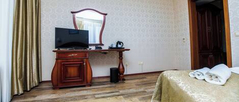 A hotel room with a wooden desk, a television, a mirror, a bed with a gold bedspread, and a wooden wardrobe.