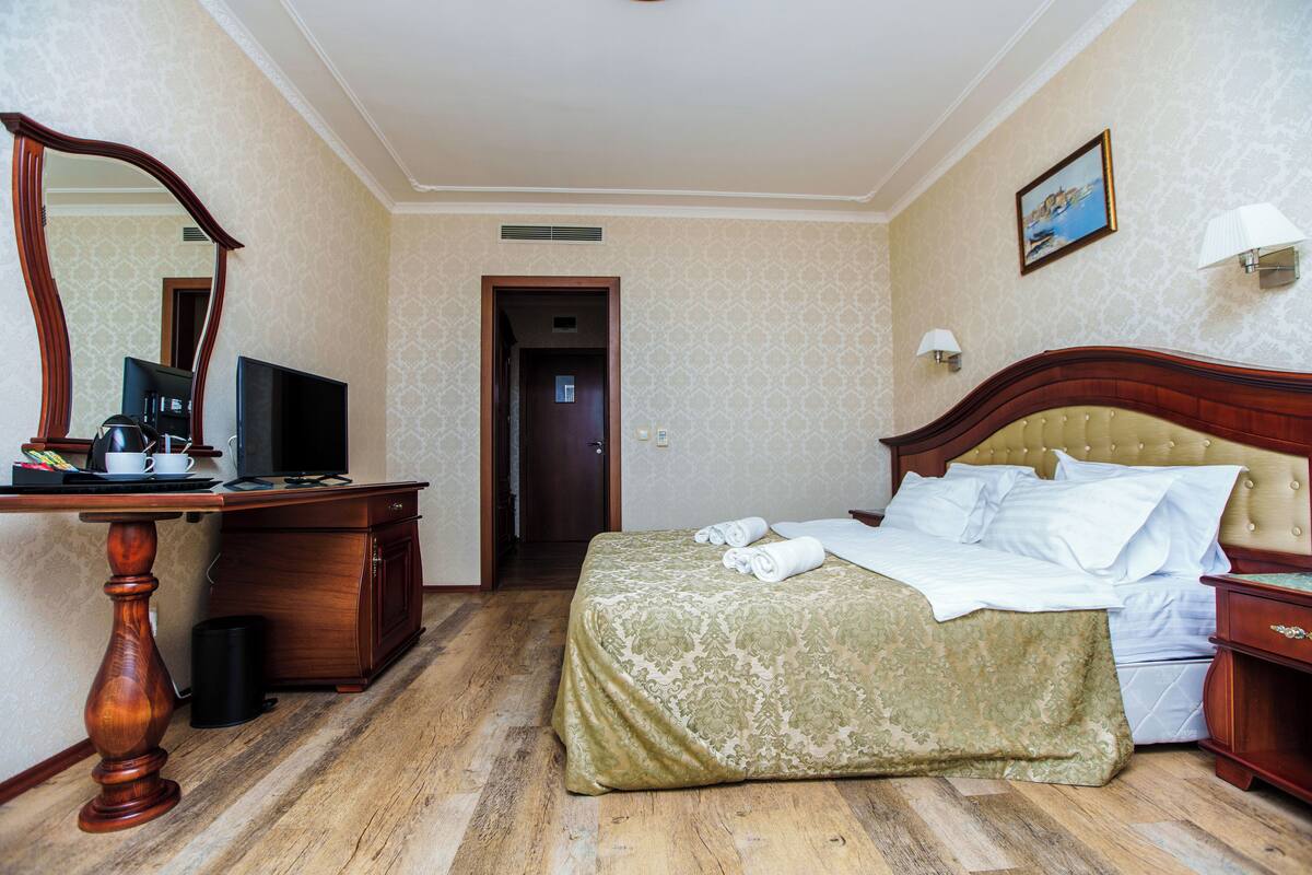 A hotel room with a wooden desk, a bed with a green bedspread, a television, and a painting on the wall.