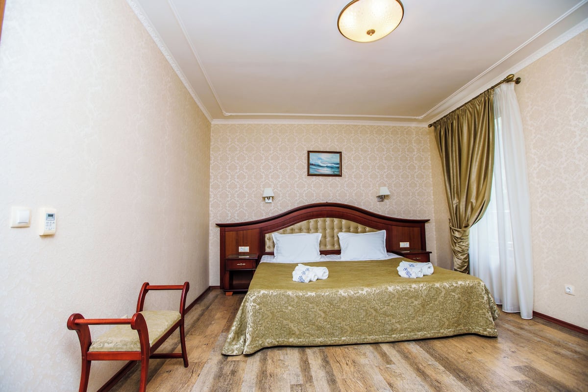 A hotel room with a wooden bed, a bedside table, a chair, and a painting on the wall.