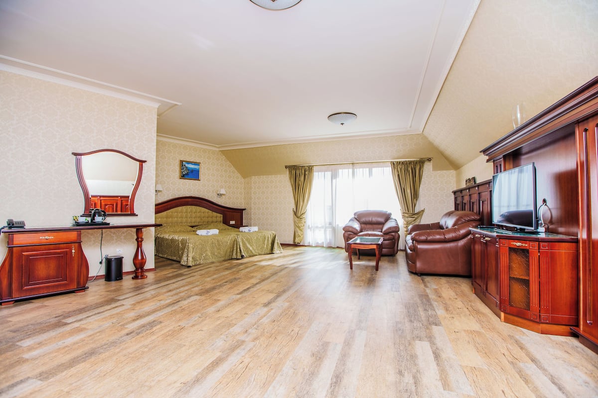 A hotel room with a wooden floor, a bed with a green bedspread, a leather armchair, a wooden desk with a television, and a large mirror.