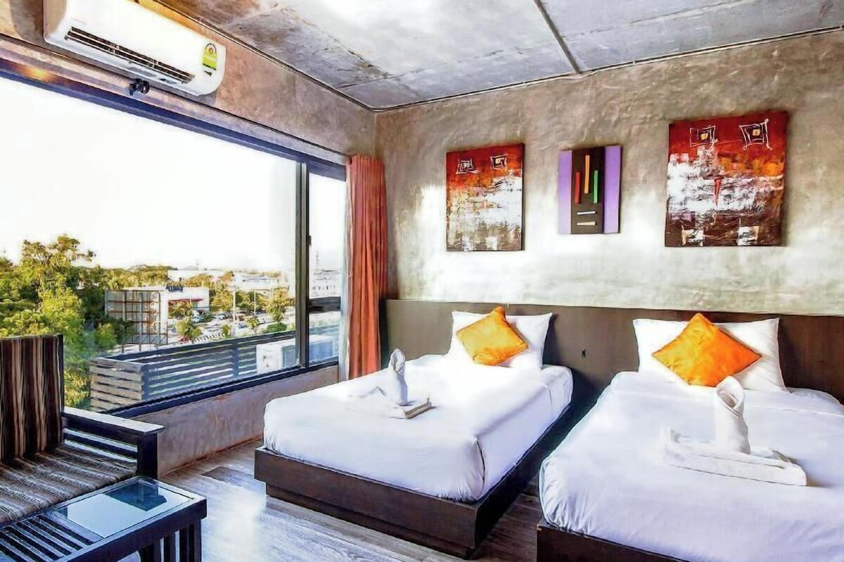 Habitación de hotel con dos camas, un sofá, un ventanal grande con vistas a la ciudad y obras de arte en las paredes.