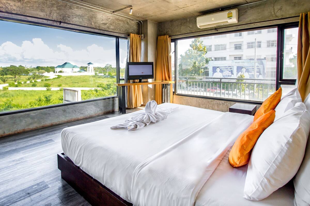 Habitación de hotel con una cama grande, televisión y vistas a zonas verdes y edificios.