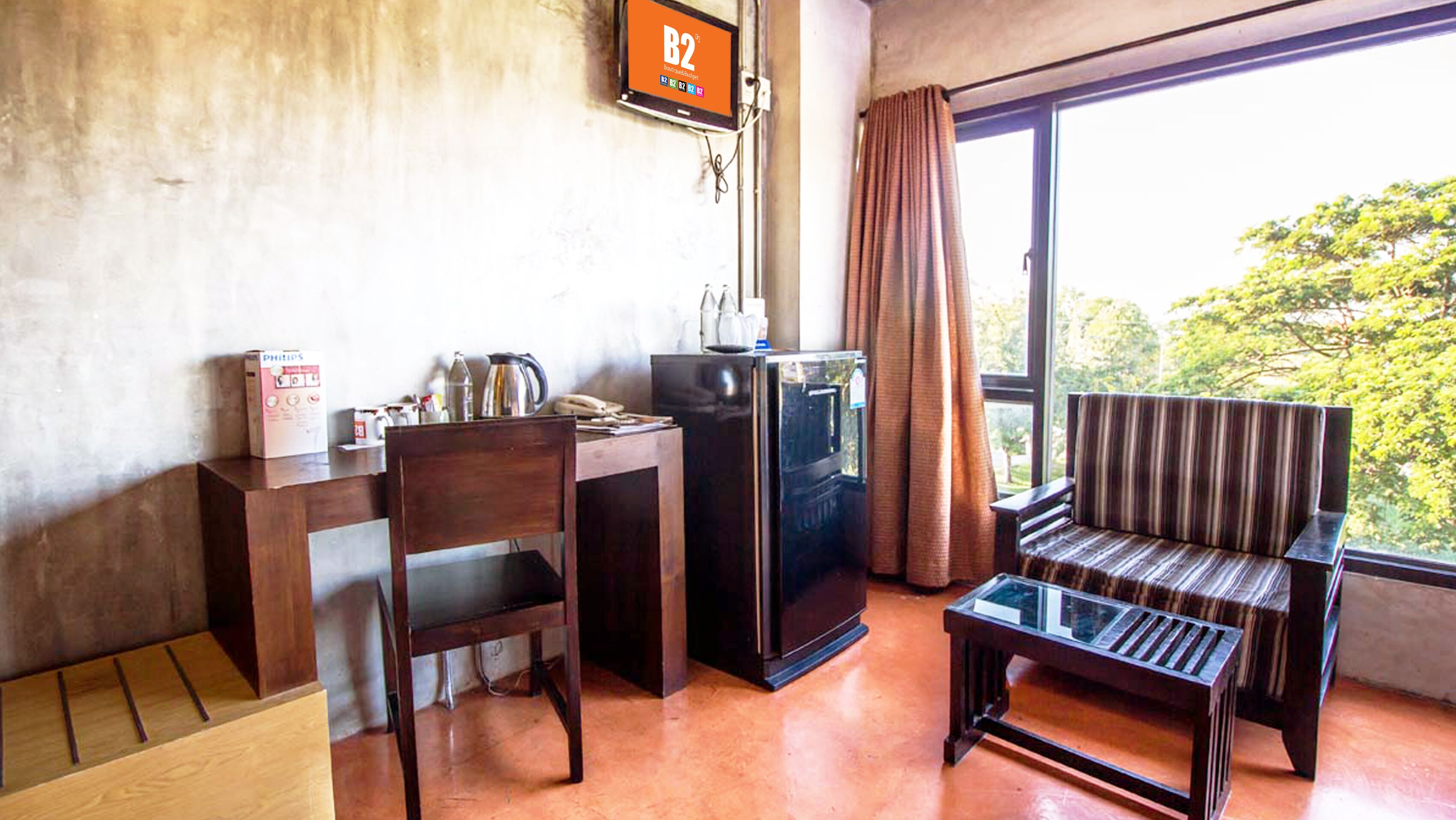 A room with a wooden desk, a chair, a small refrigerator, a sofa, and a window with curtains.