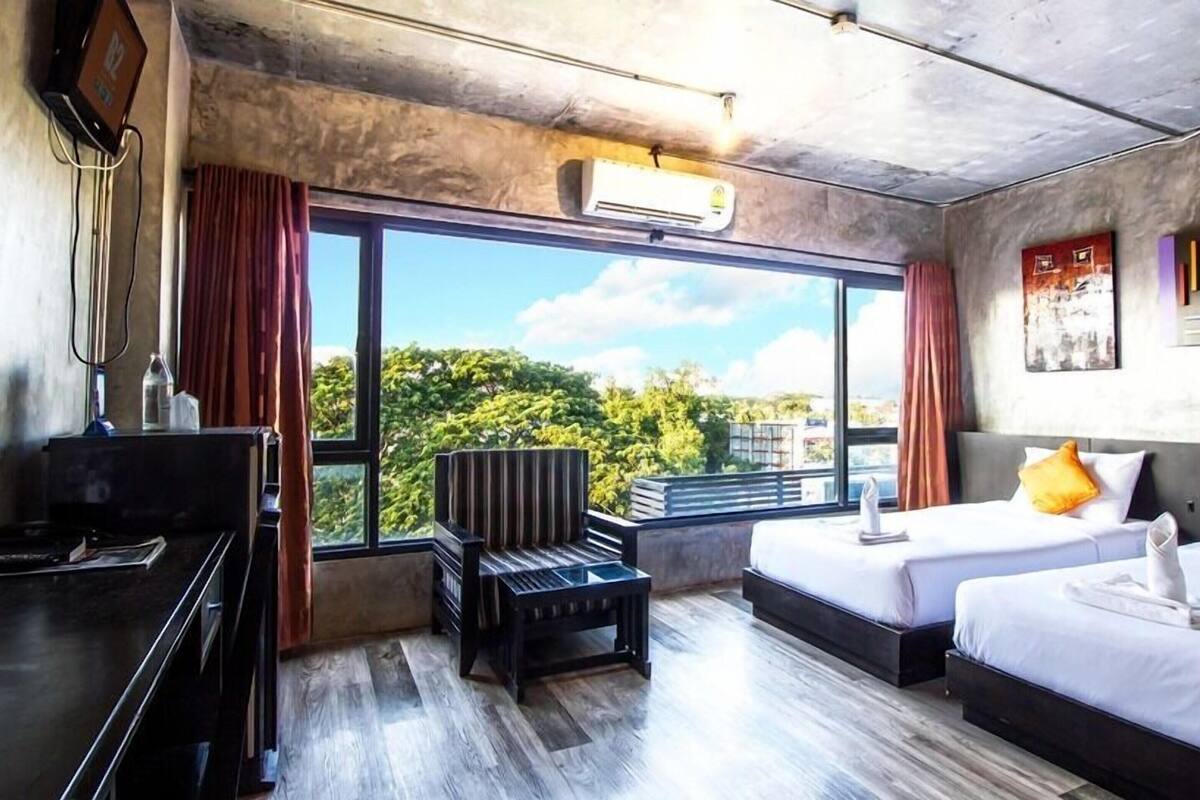 Habitación de hotel con dos camas, un escritorio, una silla y un ventanal con vistas a la vegetación.