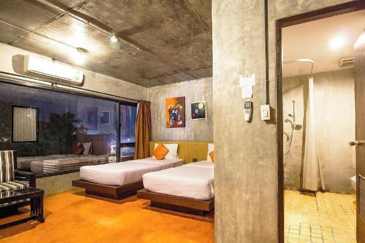 Habitación de hotel con dos camas, un sofá, un ventanal grande y un baño visible a través de una puerta abierta.