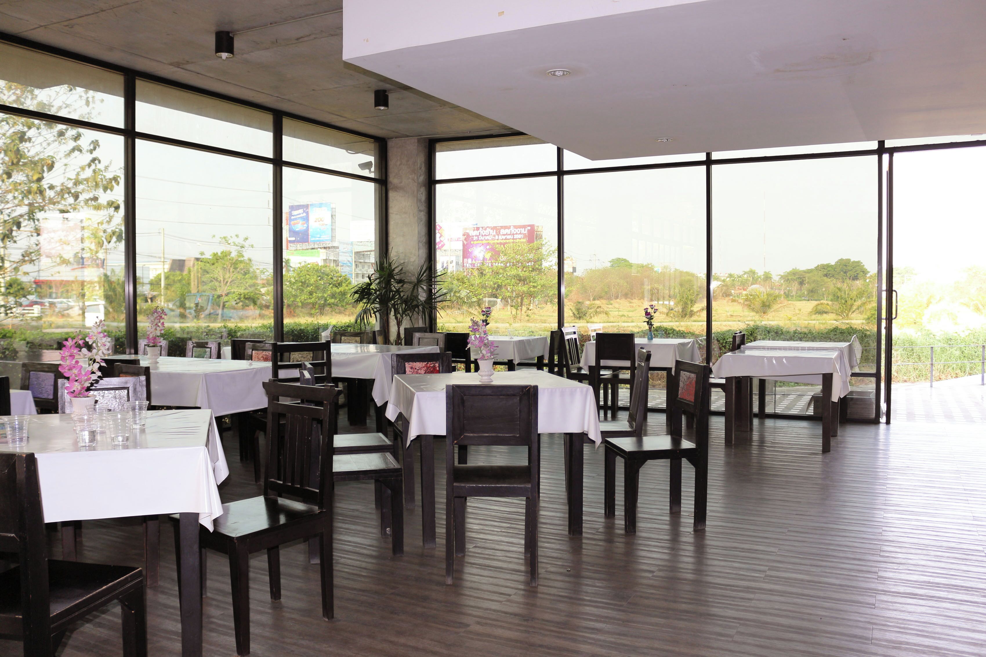 An indoor dining area with tables and chairs, large windows offering a view of greenery and buildings outside.