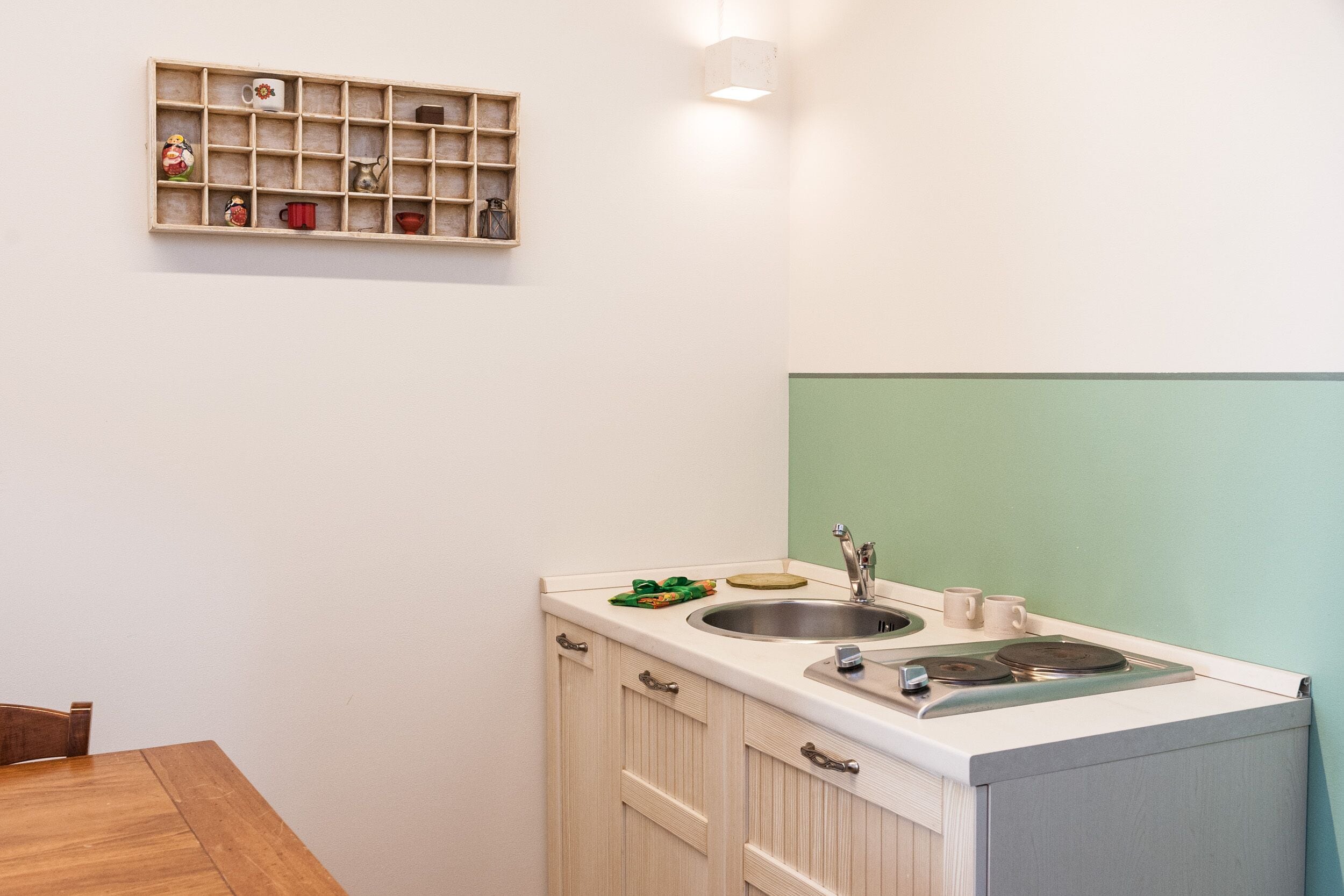 A small kitchen with a wooden table, a sink, and a shelf with various kitchen items.