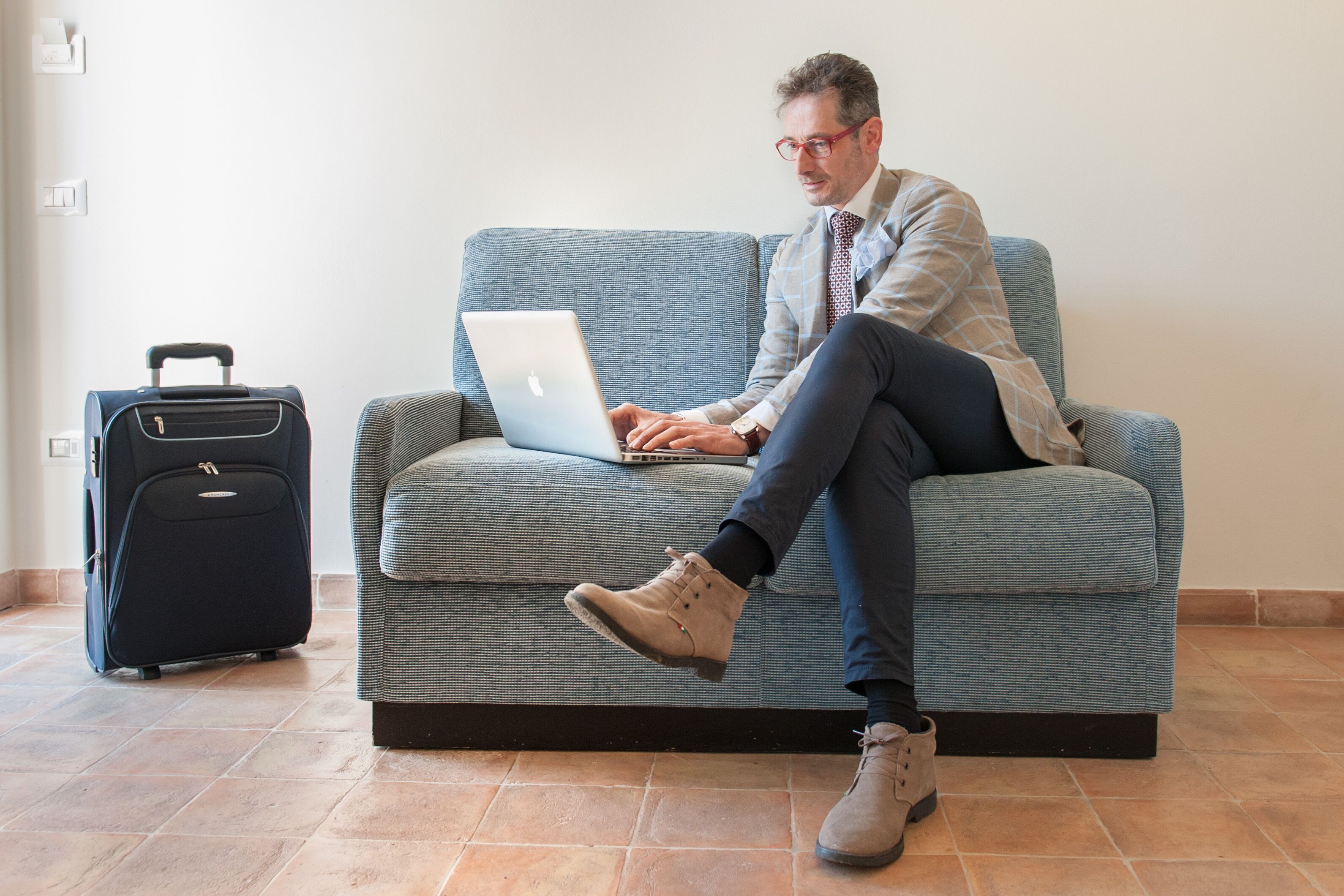 A person sitting on a sofa with a laptop, a suitcase, and a wall socket in the background.