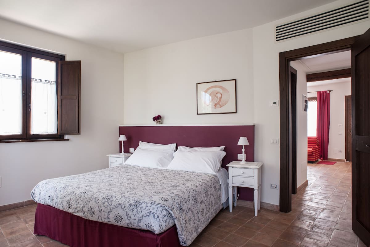 A bedroom with a bed, two bedside tables, a nightstand, a window, and a door leading to another room.