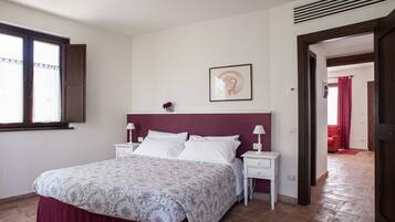 Un dormitorio con una cama, dos mesitas de noche, un velador y una ventana, además de una puerta que da a otra habitación.