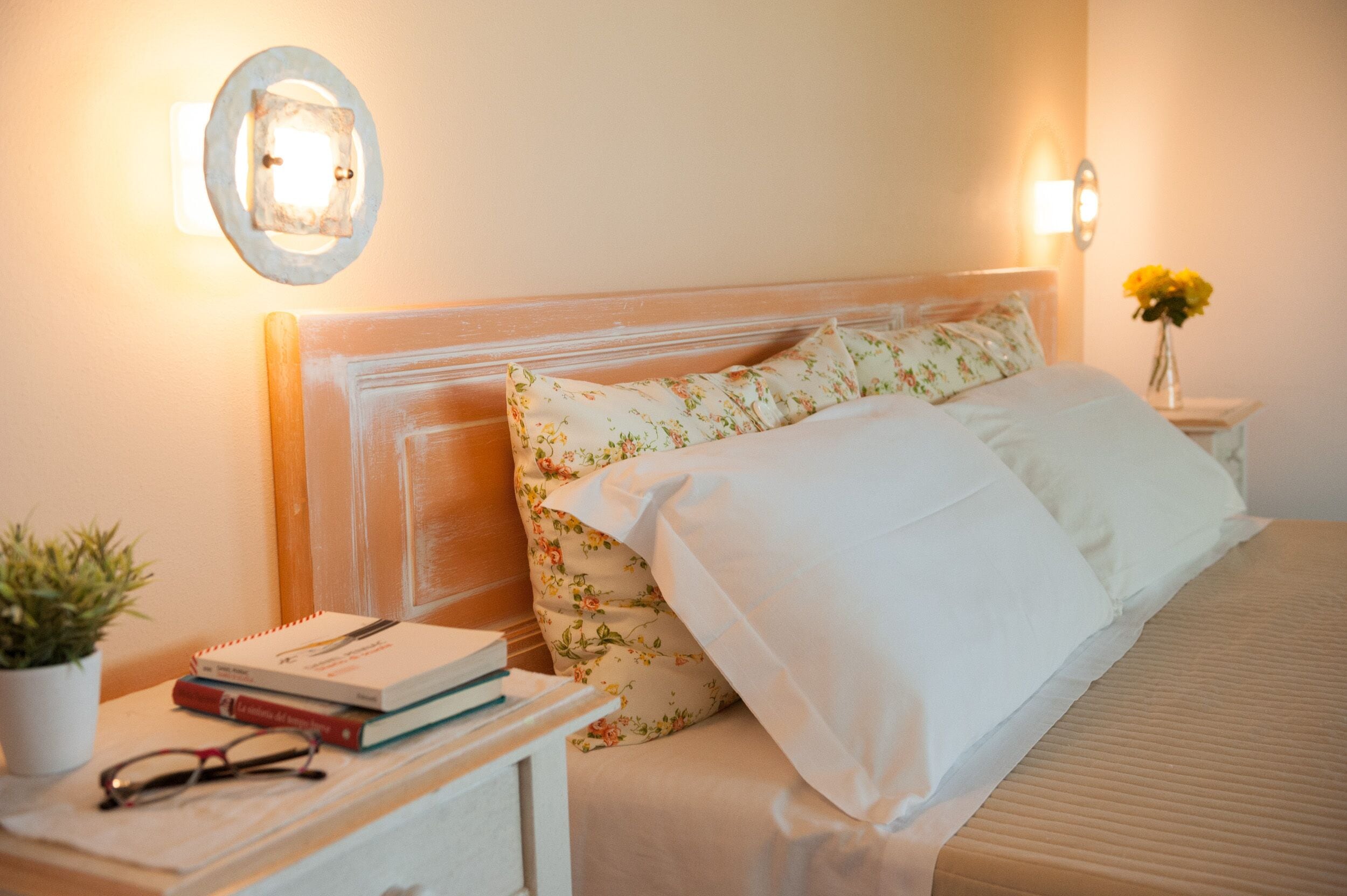 A neatly made bed with floral pillows and a white comforter, a bedside table with a book and glasses, and a wall-mounted light fixture.