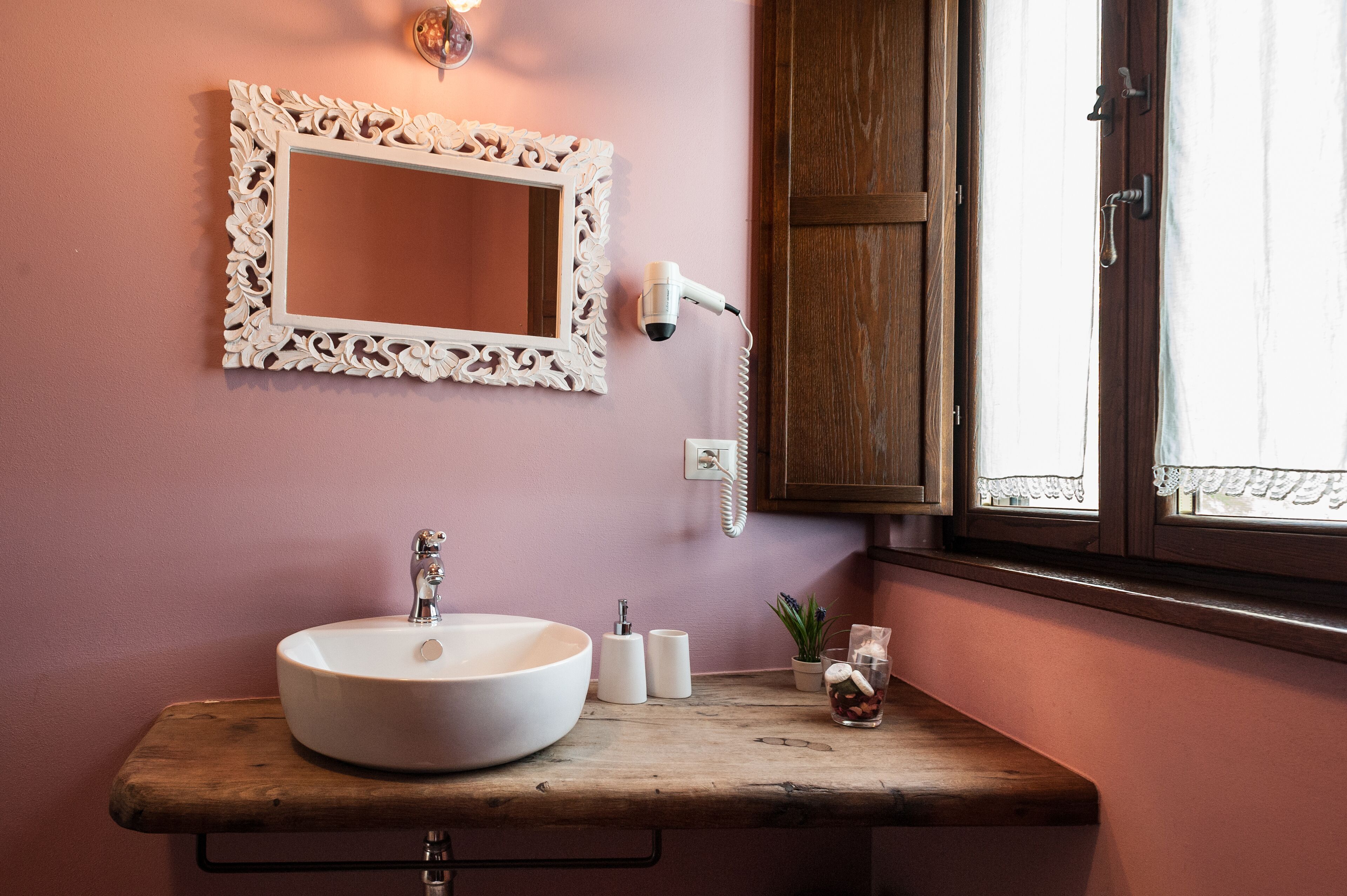 Un bagno con un lavandino bianco, uno specchio con una cornice decorata, un asciugacapelli e una finestra con tende.