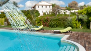A swimming pool with water cascading from a fountain, surrounded by a wooden deck and a white building in the background. - Villa Coralia (Osimo)