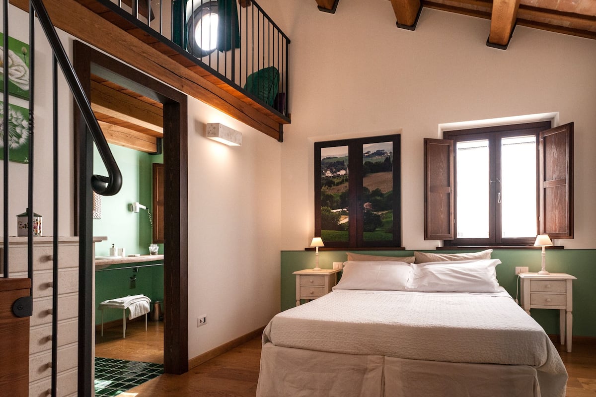 A bedroom with a bed, two bedside tables with lamps, a window with shutters, a painting on the wall, and a staircase leading to an upper level.