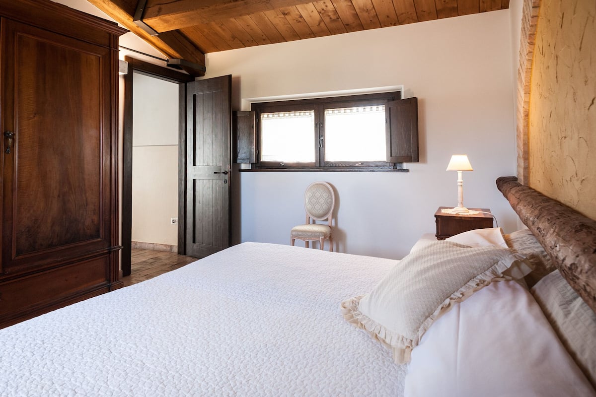 A bedroom with a large bed, a wooden wardrobe, a chair, a nightstand with a lamp, and a window with blinds.