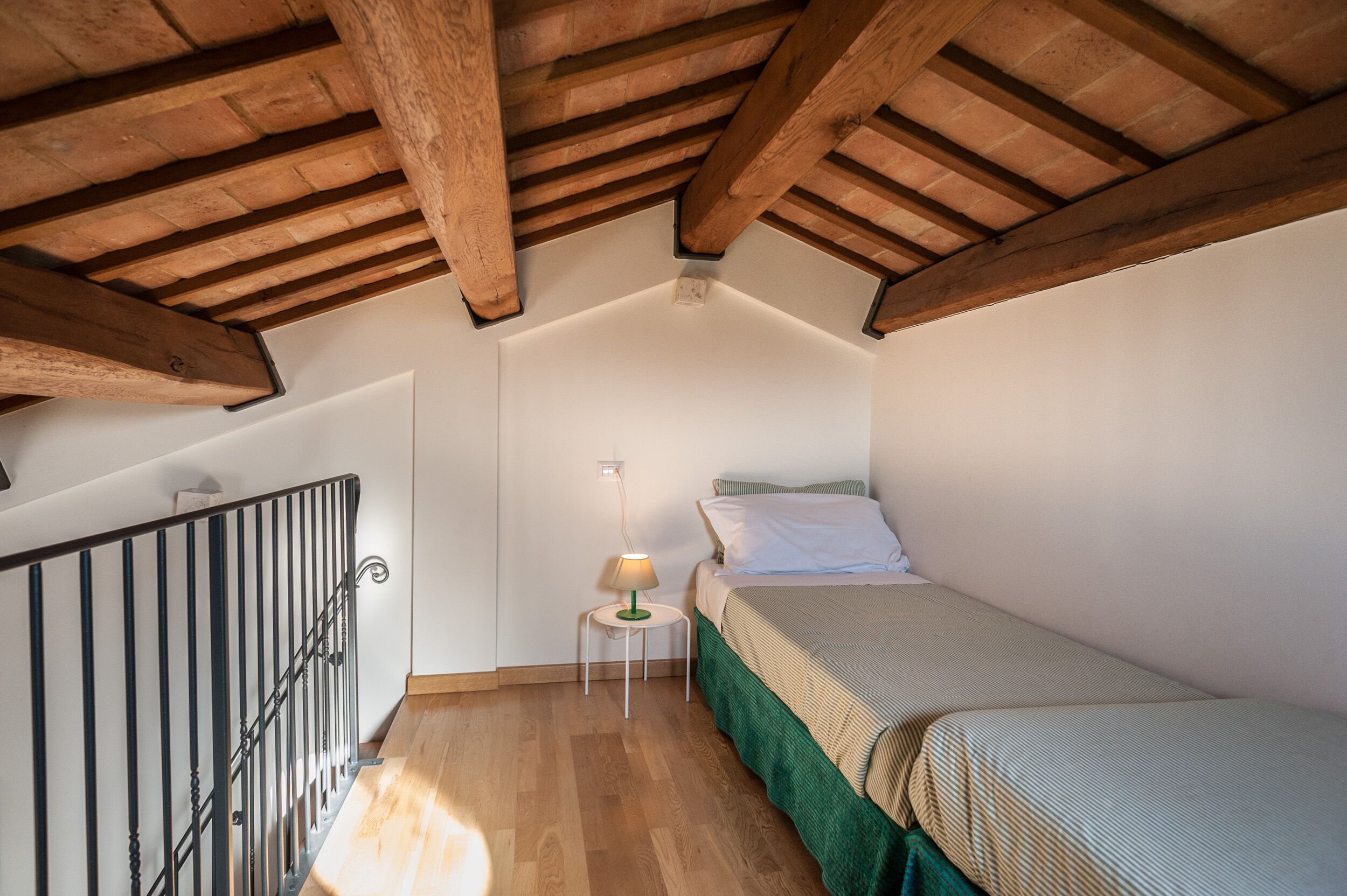 A small, cozy bedroom with a wooden ceiling, a single bed, a nightstand with a lamp, and a metal railing.