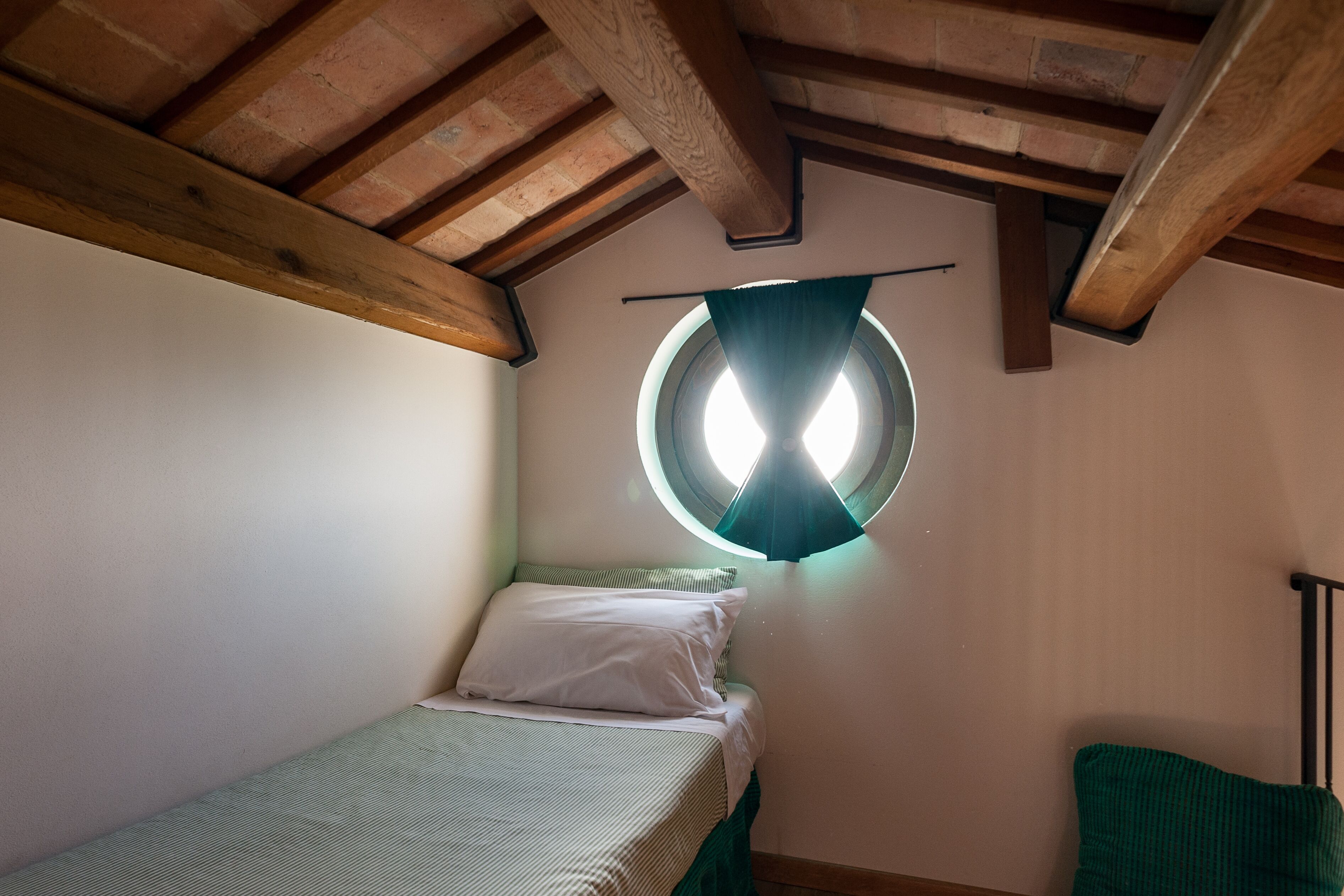 Una habitación pequeña y acogedora con una cama individual, un ventanal redondo y vigas de madera en el techo.