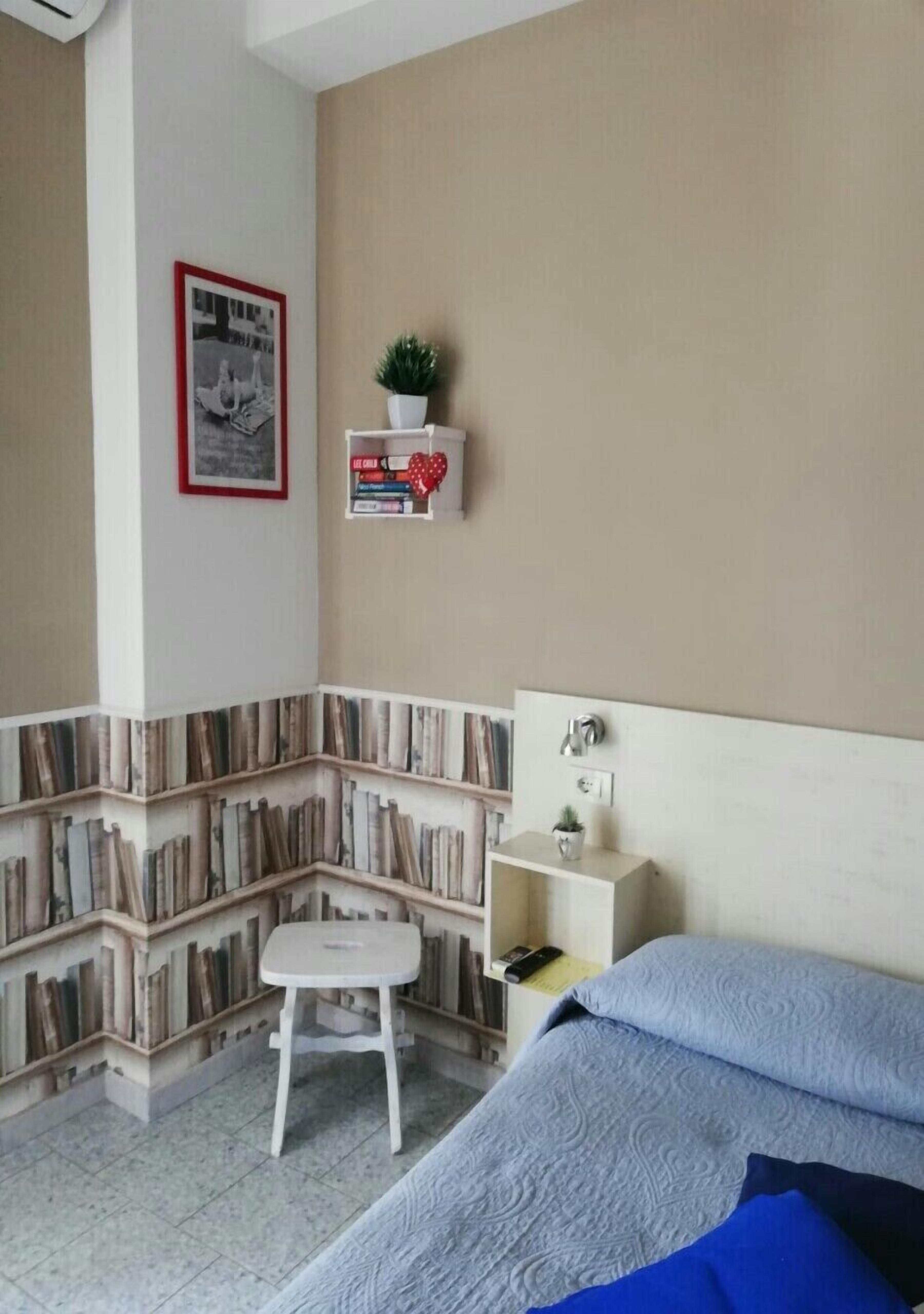 A bedroom with a bed, a small white stool, a bookshelf, and a wall-mounted shelf with a plant and books.