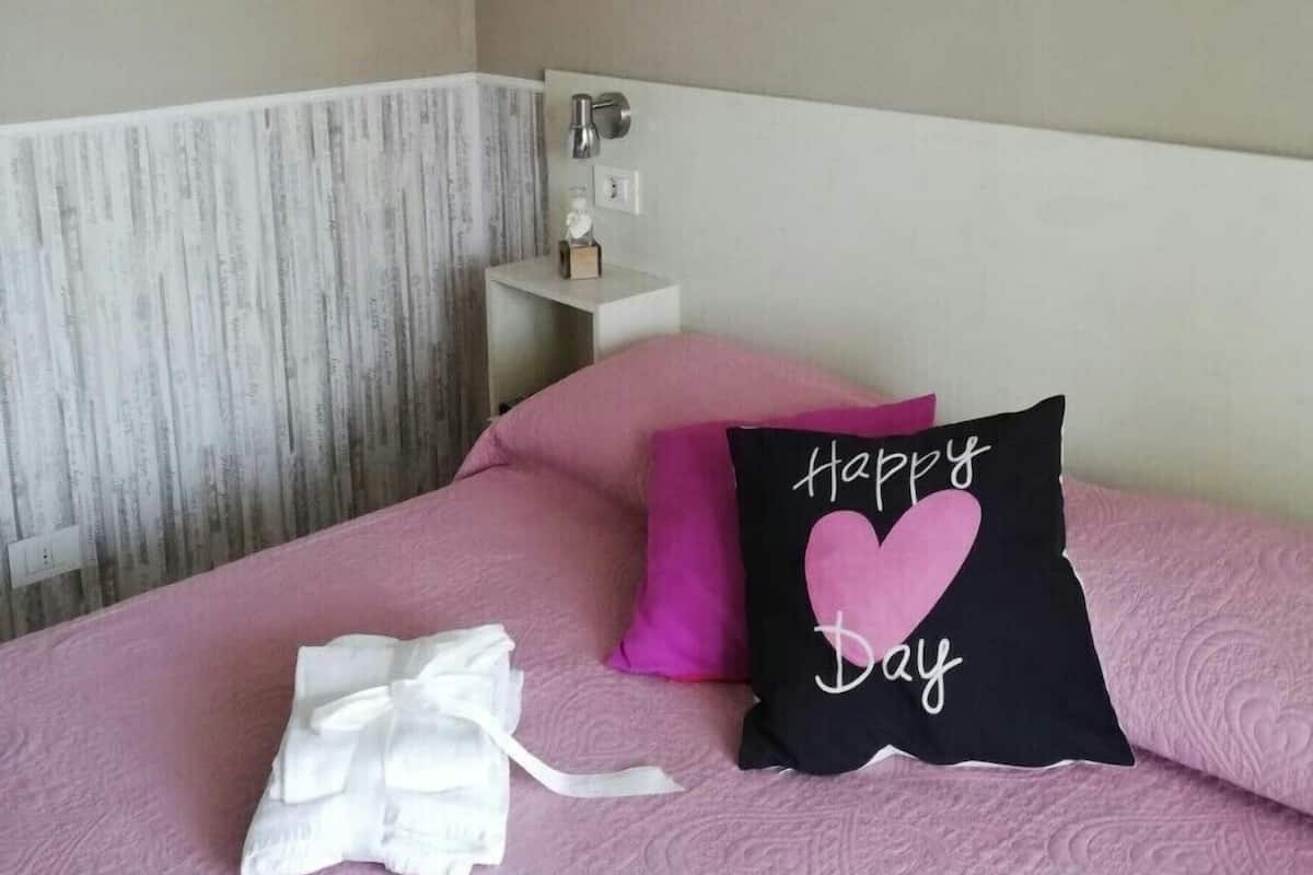 Een netjes opgemaakt bed met roze beddengoed, een kussen met de tekst 'Happy Day' en een kleine witte vaas met gedroogde bloemen aan de muur.