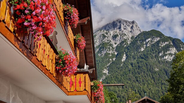 A building with wooden balconies adorned with flowers, a mountain backdrop, and a clear sky. - Gästehaus Franziska (Mittenwald)