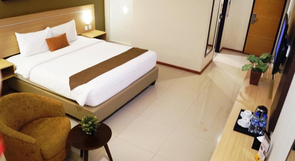 A hotel room with a large bed, a brown armchair, a small table with a plant, and a bedside table with a lamp.