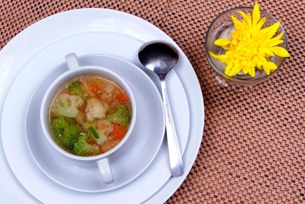 En skål suppe med ske og et glas med en gul blomst på et bord.