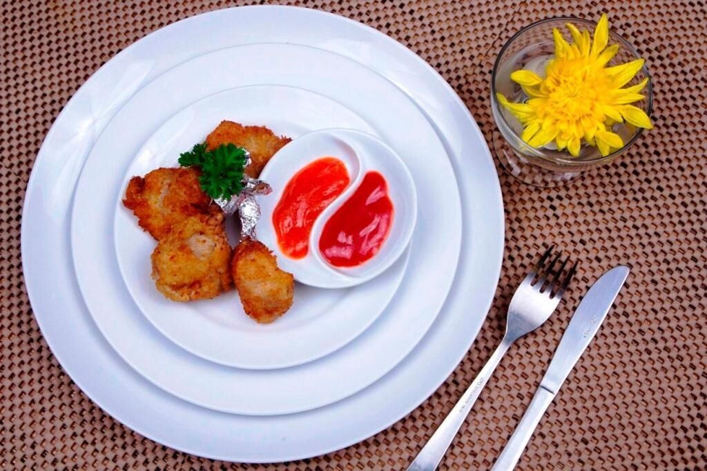 A plate with fried food and a small bowl of red sauce, a glass with yellow flowers, and cutlery on a woven surface.