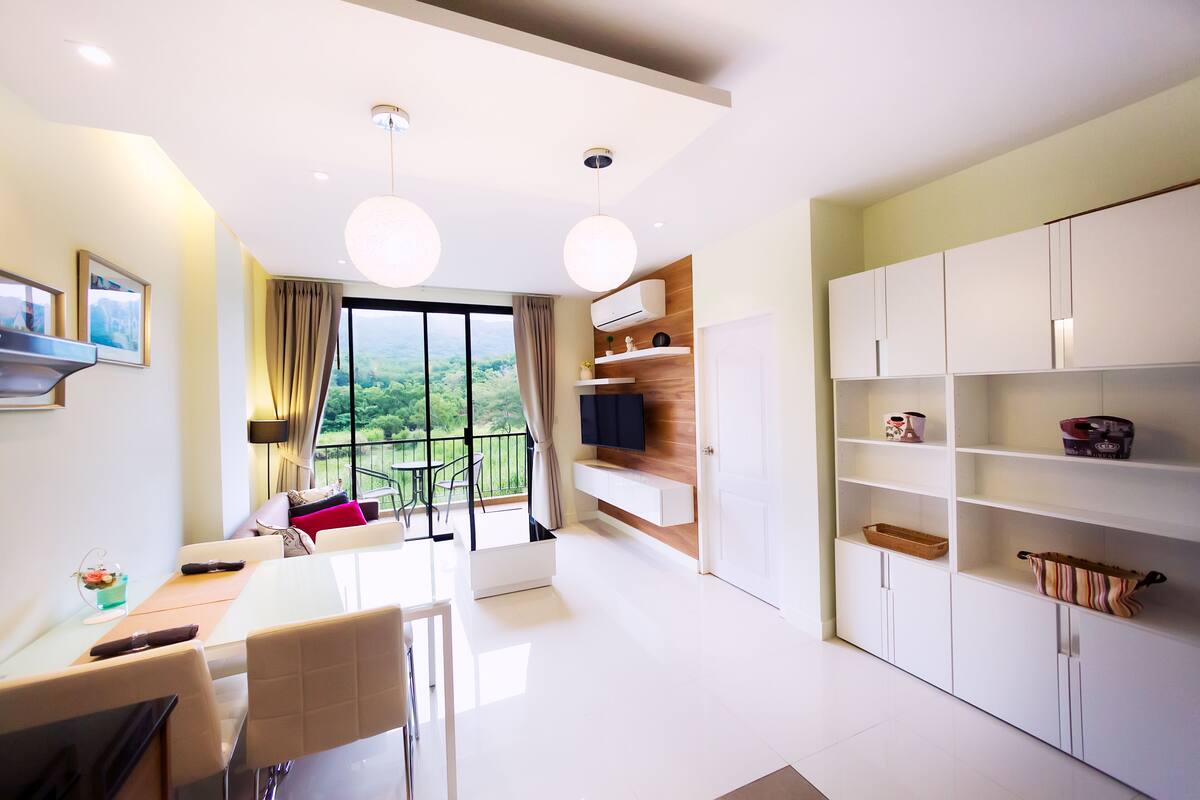 A modern living room with a dining area, large windows, and a view of greenery.