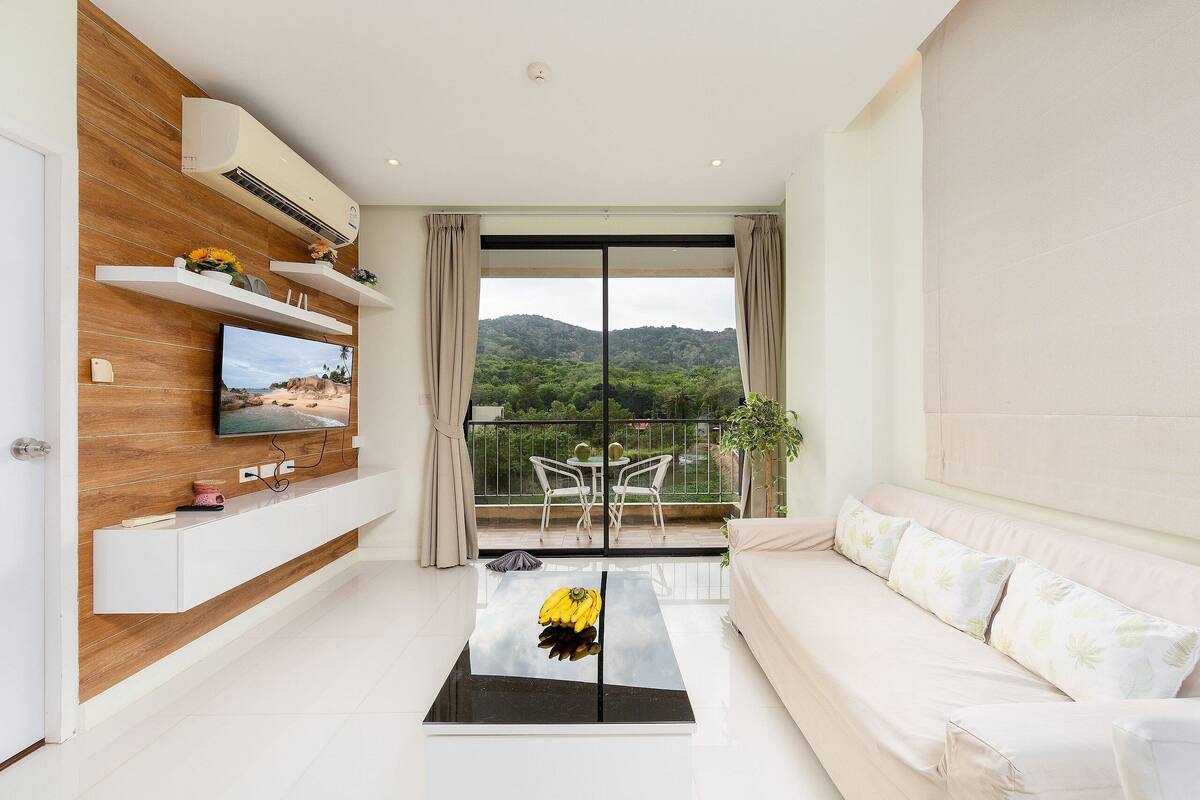 A modern living room with a flat-screen TV, a sofa, a coffee table with a banana design, and a balcony with outdoor furniture.