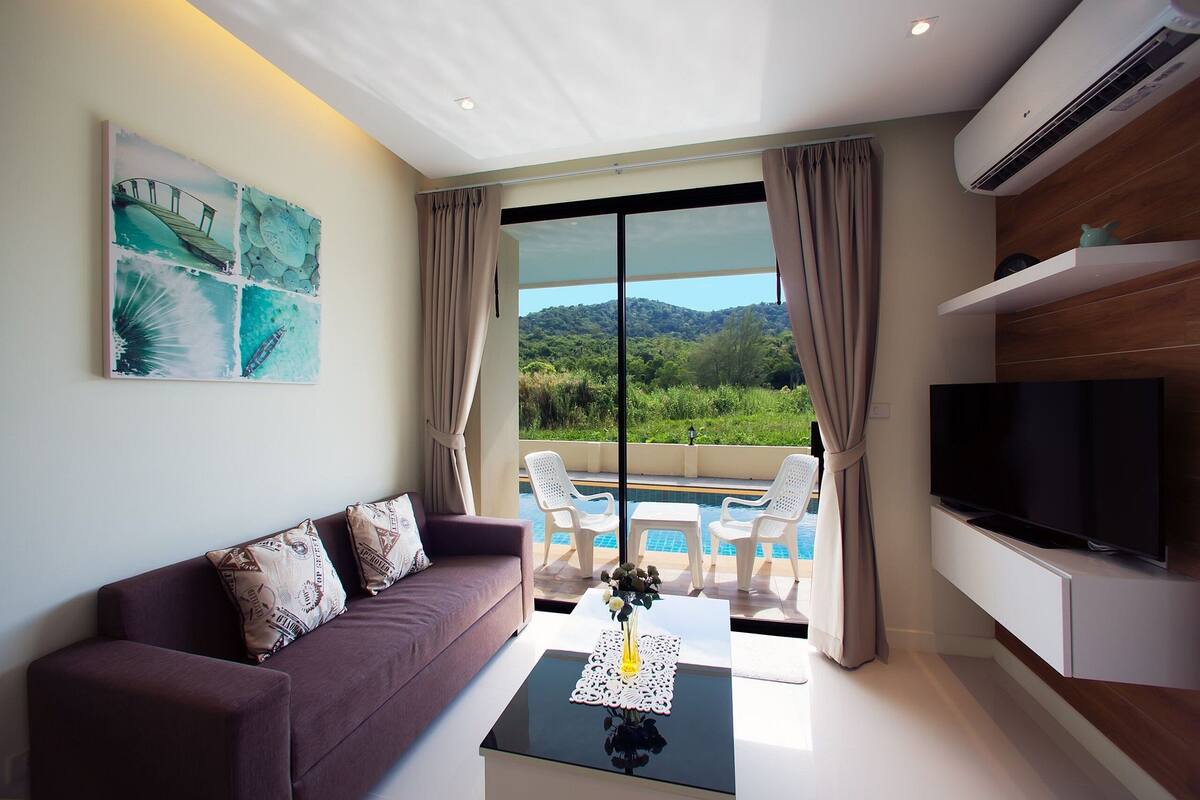 A modern living room with a sofa, a coffee table with a vase, a TV, and a pool view.