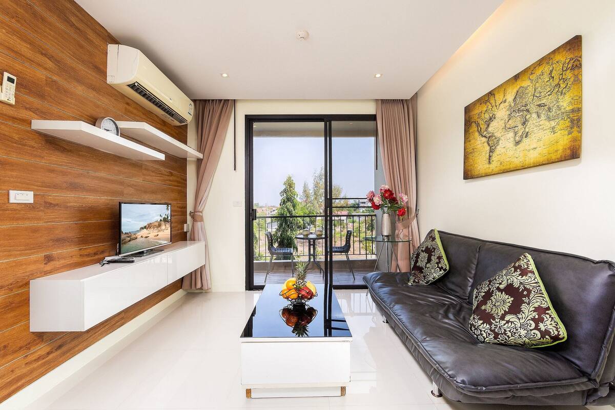 A modern living room with a flat-screen TV, a coffee table with a fruit centerpiece, a leather sofa, and a large window with a view of greenery.